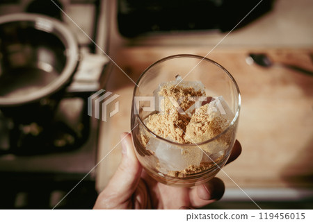 A glass filled with ice cubes and powdered Amanita muscaria mushroom on its surface, held in the hand, illuminated by natural sunlight. Preparing a homemade fly agaric elixir 119456015