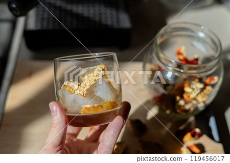 A glass filled with ice cubes and powdered Amanita muscaria mushroom on its surface, held in the hand, illuminated by natural sunlight. Preparing a homemade fly agaric elixir 119456017