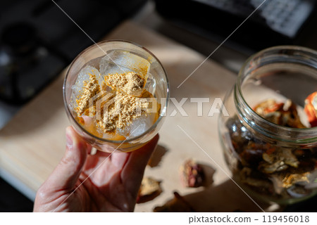 A glass filled with ice cubes and powdered Amanita muscaria mushroom on its surface, held in the hand, illuminated by natural sunlight. Preparing a homemade fly agaric elixir A glass filled with ice cubes and powdered Amanita muscaria mushroom on its surface, held in the hand, illuminated by natural sunlight. Preparing a homemade fly agaric elixir 119456018