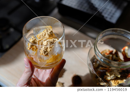 A glass filled with ice cubes and powdered Amanita muscaria mushroom on its surface, held in the hand, illuminated by natural sunlight. Preparing a homemade fly agaric elixir A glass filled with ice cubes and powdered Amanita muscaria mushroom on its surface, held in the hand, illuminated by natural sunlight. Preparing a homemade fly agaric elixir 119456019