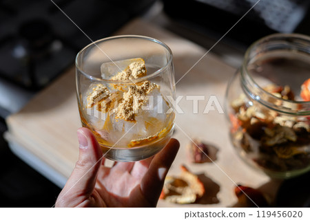 A glass filled with ice cubes and powdered Amanita muscaria mushroom on its surface, held in the hand, illuminated by natural sunlight. Preparing a homemade fly agaric elixir A glass filled with ice cubes and powdered Amanita muscaria mushroom on its surface, held in the hand, illuminated by natural sunlight. Preparing a homemade fly agaric elixir 119456020