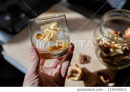 A glass filled with ice cubes and powdered Amanita muscaria mushroom on its surface, held in the hand, illuminated by natural sunlight. Preparing a homemade fly agaric elixir 119456021