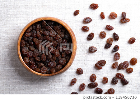 Common commercial raisins, in a wooden bowl on linen fabric. Dark-colored dried large seedless grape raisins, to be eaten raw or used in cooking, baking and brewing. Close-up, from above. Photo. Common commercial raisins, in a wooden bowl on linen fabric. Dark-colored dried large seedless grape raisins, to be eaten raw or used in cooking, baking and brewing. Close-up, from above. Photo. 119456730
