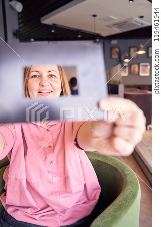 Smiling woman in pink shirt framing face with cardboard cutout in cozy cafe Smiling woman in pink shirt framing face with cardboard cutout in cozy cafe 119461944