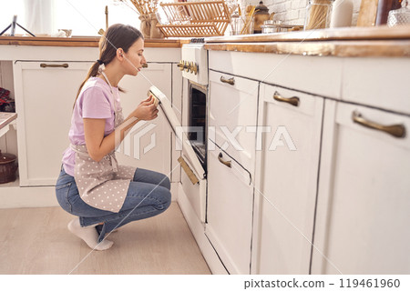 Woman baking in modern kitchen with apron, checking food in oven, home cooking Woman baking in modern kitchen with apron, checking food in oven, home cooking 119461960