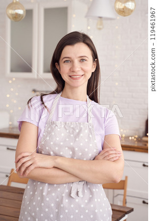 Smiling woman in polka dot apron standing in cozy kitchen with white brick walls Smiling woman in polka dot apron standing in cozy kitchen with white brick walls 119461967