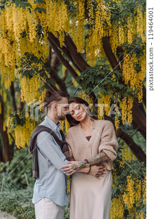 Amazing couple hugging under tree with yellow tree. Lovers couple hugging in a laburnum tree. Couple lovers hugging in the park. Girl with tattoos. A girl and a boy kiss. Amazing couple hugging under tree with yellow tree. Lovers couple hugging in a laburnum tree. Couple lovers hugging in the park. Girl with tattoos. A girl and a boy kiss. 119464001