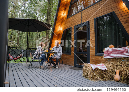 Couple enjoying meal on wooden terrace of A-frame cabin in the forest, creating a cozy autumn vibe Couple enjoying meal on wooden terrace of A-frame cabin in the forest, creating a cozy autumn vibe 119464560