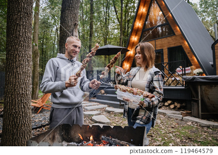 Couple preparing skewers for barbecue over open fire at cabin, enjoying autumn grilling together 119464579