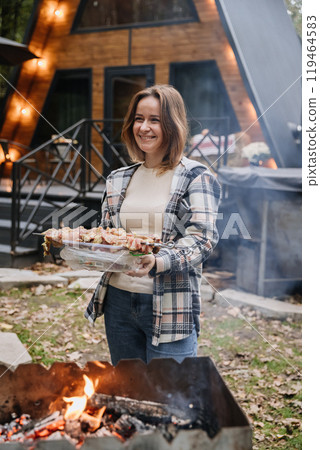 Woman preparing skewers for barbecue on an open fire, enjoying outdoor cooking with a smile 119464583