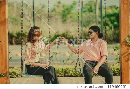 Young couple in love sitting on a swing making a heart shape. Happy couple in love on a park swing making a heart shape Young couple in love sitting on a swing making a heart shape. Happy couple in love on a park swing making a heart shape 119466961