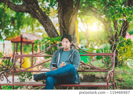 Handsome man sitting on a bench checking cell phone at sunset. Young man sitting on a park bench texting on cell phone 119466987