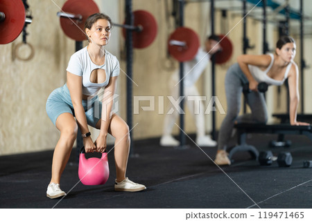 Young girl doing exercises with kettlebell near other people in crossfit gym 119471465