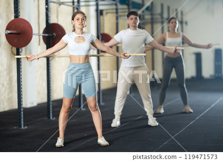 Slender girl, together with other athletes, does a warm-up using a long stick in gym Slender girl, together with other athletes, does a warm-up using a long stick in gym 119471573