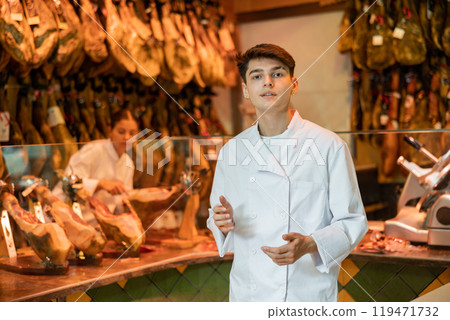 Young guy seller in white jacket selling spanish jamon in butcher shop 119471732