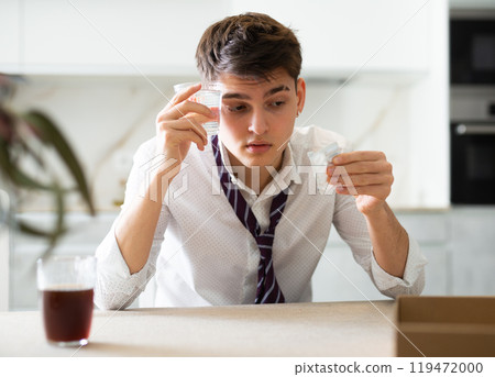Sick young man with hangover drinks pills while sitting at table Sick young man with hangover drinks pills while sitting at table 119472000