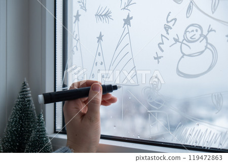 Sustainable responsible holiday decor. Female hand painting Christmas tree painted on window glass indoor at home. Christmas winter decoration. White pattern. Snow art tracery on season. Diy 119472863
