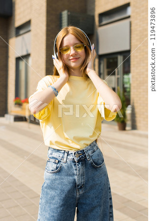 Happy teenager girl in a yellow shirt and blue jeans is wearing headphones and smiling. She is standing on a sidewalk in front of a building 119473896