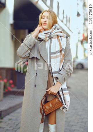 A woman dressed in a chic overcoat and plaid scarf stands on a lively street, showcasing her fashionable urban look 119475619