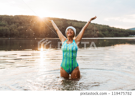 Cold water swimming for elderly women. Senior sporty women standing in lake during cold evening, arms in the air. Cold water swimming for elderly women. Senior sporty women standing in lake during cold evening, arms in the air. 119475782
