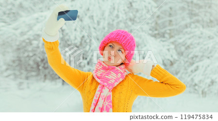 Happy cheerful smiling young woman taking selfie with smartphone in hat, sweater in snowy park 119475834