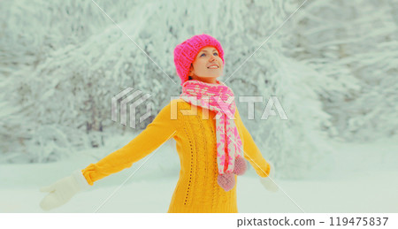Happy cheerful smiling young woman in colorful knitted hat, enjoys weather in winter snowy park 119475837