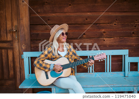 Beautiful older women is sitting in front of her cabin, playing on guitar. Beautiful older women is sitting in front of her cabin, playing on guitar. 119475840