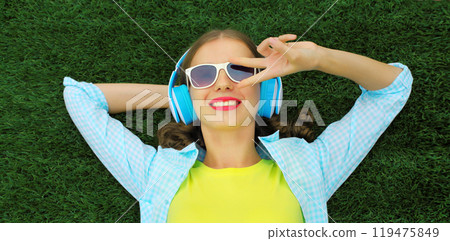 Happy smiling young woman listening to music in headphones while lying on grass in summer park 119475849