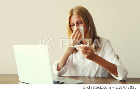 Sick woman sneezing blow nose, measuring body temperature with thermometer working with laptop Sick woman sneezing blow nose, measuring body temperature with thermometer working with laptop 119475853