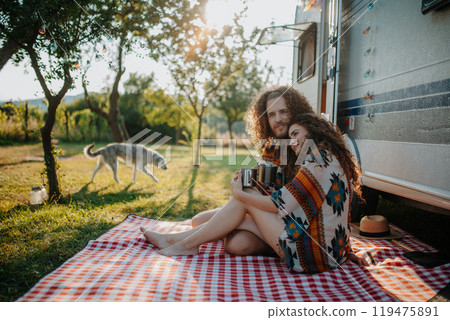 Couple sitting by caravan, enjoying peaceful moment, drinking coffee of herbal tea. Camping trip for young people. 119475891