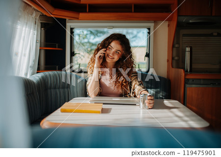 Young beautiful woman is on a camping trip in nature, sitting in her caravan making phone call. Caravan as homeoffice, working remotely. Young beautiful woman is on a camping trip in nature, sitting in her caravan making phone call. Caravan as homeoffice, working remotely. 119475901