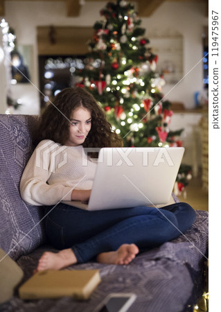 Young woman is shopping for Christmas gifts online using her laptop, sitting on the couch in living room with Christmas decorations. Young woman is shopping for Christmas gifts online using her laptop, sitting on the couch in living room with Christmas decorations. 119475967