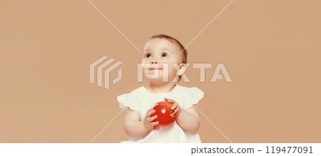 Portrait of cute baby holding red apple fruit sitting looking up on brown studio background Portrait of cute baby holding red apple fruit sitting looking up on brown studio background 119477091