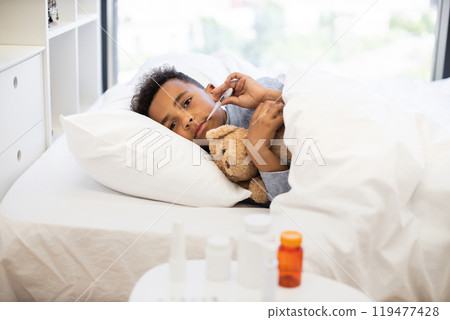 African boy with flu lying in bed holding thermometer and teddy bear 119477428