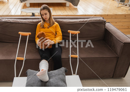 Young girl with broken leg cast sitting on sofa. Sad little kid hugging her teddy bear recovering from broken leg at home. Girl using cast and crutches. Accident injury fracture treatment foot bandage 119479061