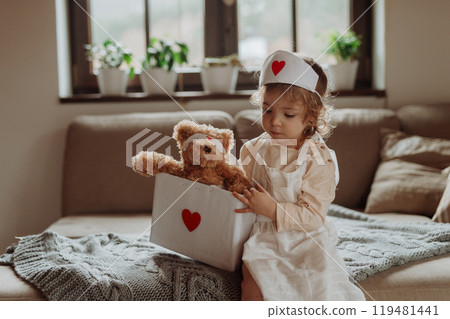 Girl in doctor uniform is playing hospital. Little girl as nurse taking care of sick teddy bear. 119481441