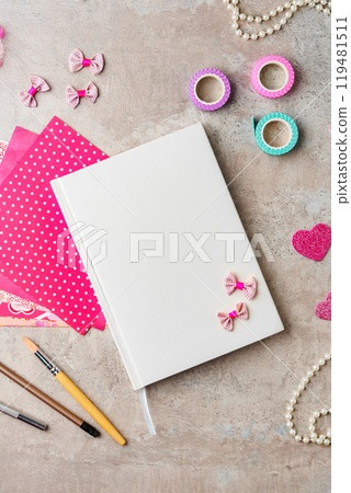 A collection of crafting materials arranged on a textured surface with a blank notebook, decorative paper, and various embellishments A collection of crafting materials arranged on a textured surface with a blank notebook, decorative paper, and various embellishments 119481511