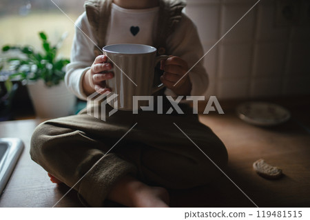 Adorable little girl is drinking tea. The sick girl with cold is sitting on kitchen counter with mug in hand. 119481515