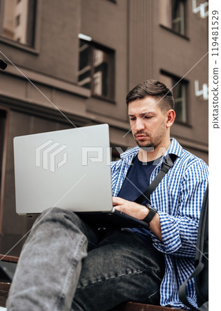 Young man working on a laptop while seated on a bench outside 119481529