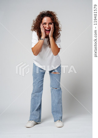 A woman in a white shirt and ripped jeans posing positively with a playful expression in a light studio setting 119481579