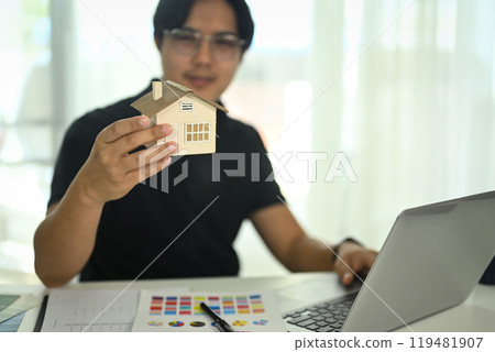 Man holding a small model house while reviewing details of the model at desk with color samples and material swatches 119481907