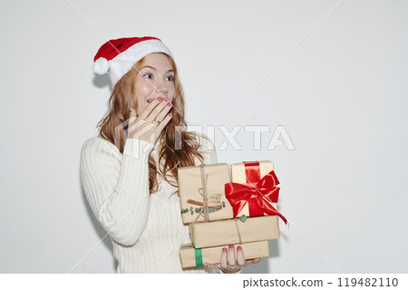 Surprised Woman Holding Christmas Gifts Wearing Santa Hat 119482110