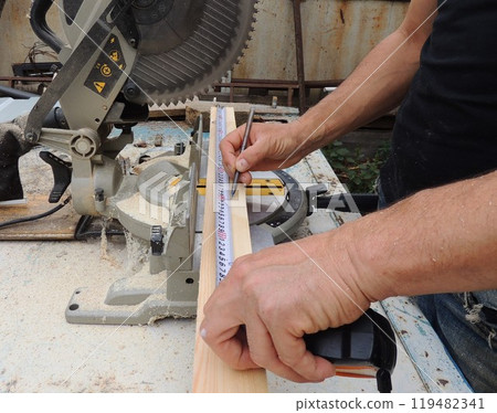 male hands with tape measure and pencil measure length of wooden plank on workbench with miter saw, marking material before processing with circular saw 119482341