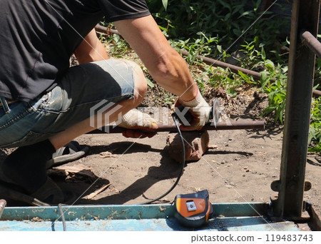 a man with gloves sawing off a part of a narrow iron pipe from behind with a grinder outdoors, working with iron, processing metal products at home with a hand tool a man with gloves sawing off a part of a narrow iron pipe from behind with a grinder outdoors, working with iron, processing metal products at home with a hand tool 119483743