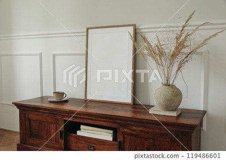 Modern autumn interior still life. Scandinavian interior design. Blank picture mock up poster frame. Mahogany wooden dresser sideboard. Dry grass in clay vase. Cup of tea, coffee. White wall 119486601