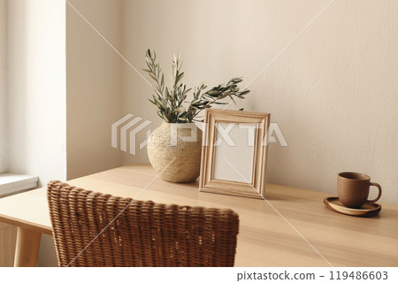 Elegant artistic mockup template. Black vertical photo frame on wooden table desk. Vase with olive tree branches, cup of tea, coffee. Poster display. Blurred rattan chair. Summer Mediterranean design. 119486603