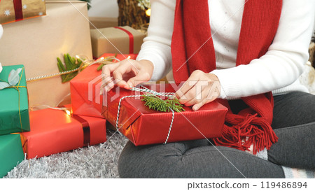 Close up woman hands wrap present gift in cozy living room. Woman wrapping christmas gift box under xmas tree. Asian woman decor bauble ball christmas tree in xmas holiday winter seasonal celebration Close up woman hands wrap present gift in cozy living room. Woman wrapping christmas gift box under xmas tree. Asian woman decor bauble ball christmas tree in xmas holiday winter seasonal celebration 119486894