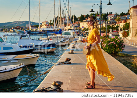 Woman is walking alone on sunny promenade with palm trees and flowers. Female tourist strolls along marina at sunset in coastal town Woman is walking alone on sunny promenade with palm trees and flowers. Female tourist strolls along marina at sunset in coastal town 119487392