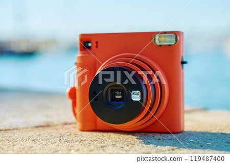 Vintage orange instant camera on rock by seaside with blurred coastal town in background. Summer travel concept. Equipment for capturing moments 119487400