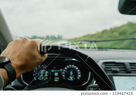 Driver hand gripping steering wheel with view of countryside road ahead through windshield. Man driving car in rainy day. Inside view of modern vehicle interior Driver hand gripping steering wheel with view of countryside road ahead through windshield. Man driving car in rainy day. Inside view of modern vehicle interior 119487428